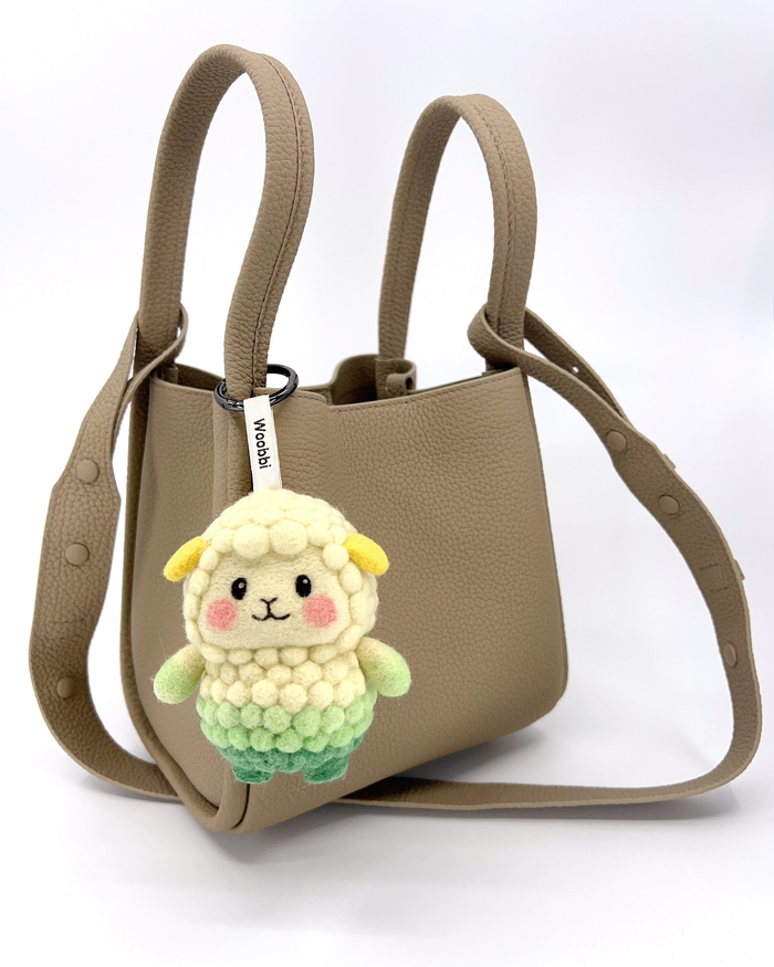 A taupe, textured leather mini tote bag with two top handles and a shoulder strap is shown against a plain background. Hanging from the front handle is a small, needle-felted sheep keychain. The sheep has a creamy white bubbly wool body and head, a light green lower body, small yellow ears, and pink cheeks. It is attached with a white tag that reads "Woobbi".