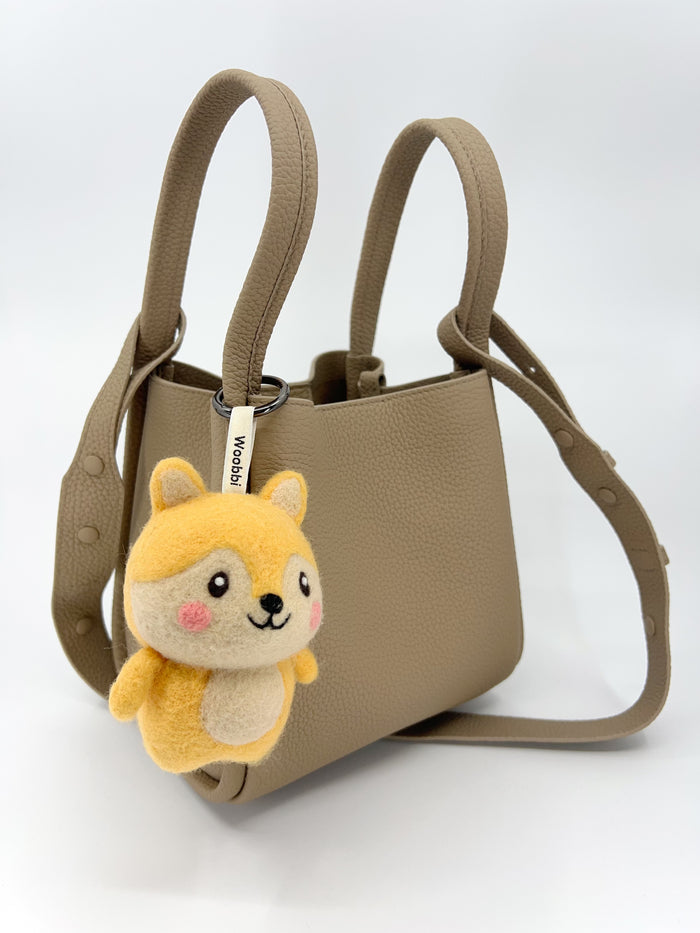 A tan leather mini tote bag shown against a plain white background, featuring a handmade Woobbi fox keychain hanging from the front handle. The fox charm is made of yellow and cream wool felt with pink cheeks and a smiling face. A white tag labeled 'Woobbi' connects the charm to the bag's hardware.