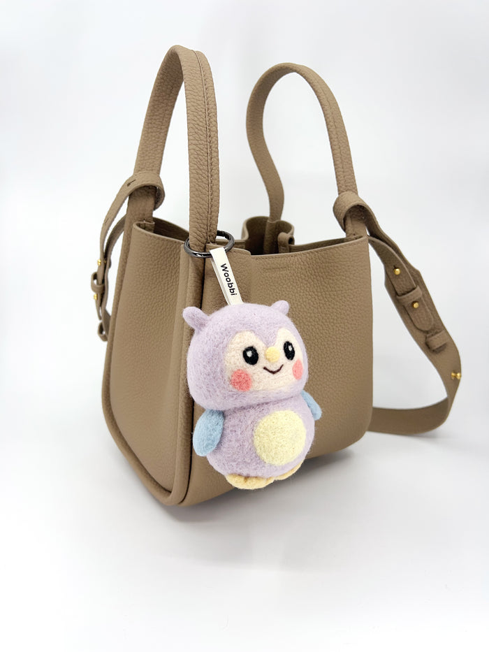 Handcrafted needle-felted wool bag charm featuring a cute purple owl design, styled on a beige leather bucket bag to show size and texture.