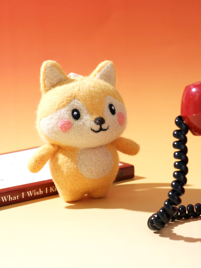 Handcrafted needle-felted yellow fox bag charm standing next to a vintage red rotary phone, styled on a warm orange background.