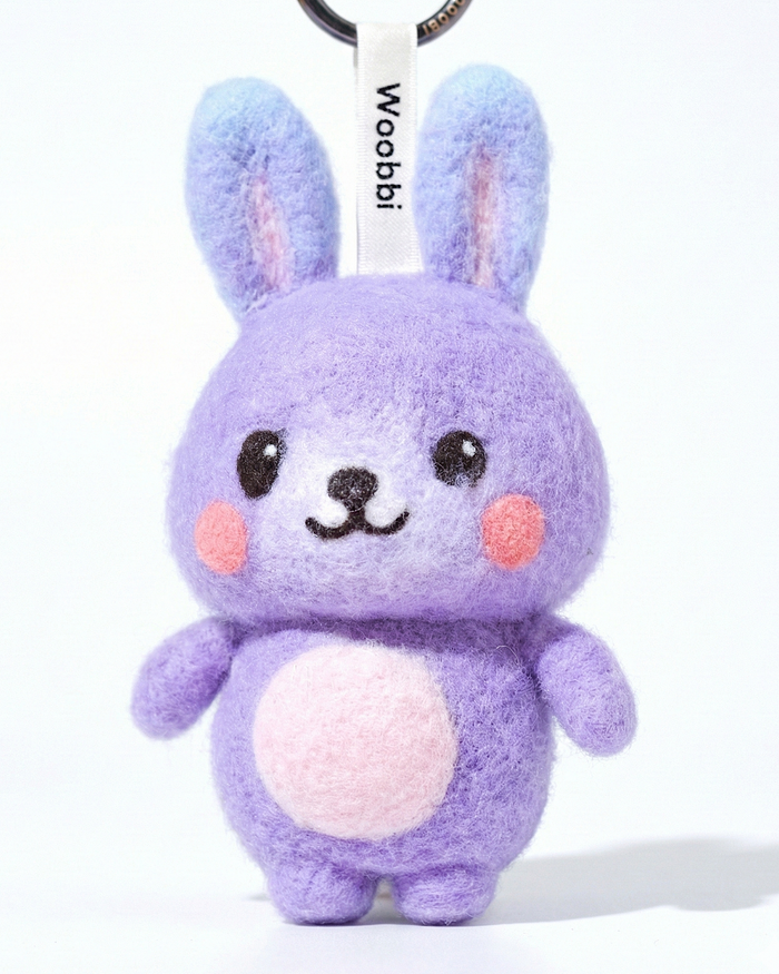 Front view of a handmade Woobbi bunny keychain, crafted from purple bubbly wool felt. It has small yellow ears, rosy pink cheeks, and a smiling face with black eyes and nose. A small tag with the brand name 'Woobbi' is attached to the top. It is set against a plain white background.