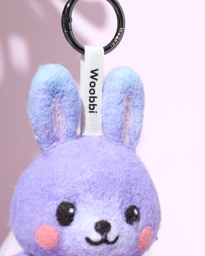 Close-up of black metal keychain hardware and white woven logo tag attached to a purple wool bunny charm with gradient blue ears.