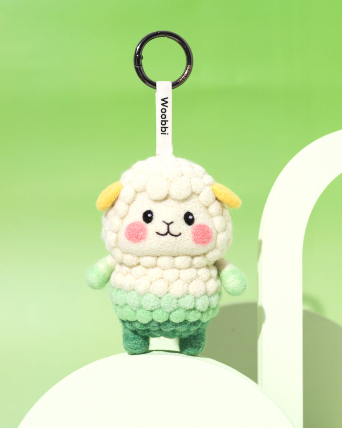 Front view of white and green needle-felted sheep keychain standing on a modern green arch, designed in Austin with sustainable Merino wool.
