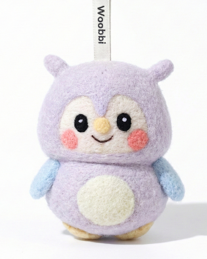 Front view of a handmade Woobbi owl keychain, crafted from soft lavender wool felt. The owl features large black eyes, rosy pink cheeks, a small yellow beak, and a cream-colored belly patch. It has light blue wings and is attached to a black metal keyring with a white tag labeled 'Woobbi'. The background is plain white with soft studio lighting.