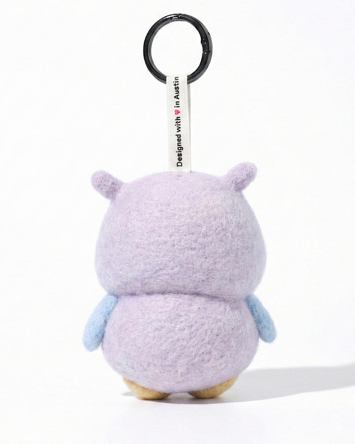 Back view of a handmade Woobbi owl keychain, showing its rounded lavender wool felt body and small pointed ears. The keychain features light blue wings on the sides and a white tag attached to the top that reads 'Designed with ❤️ in Austin'. A round black metal keyring is connected to the tag. The product is set against a pure white background.