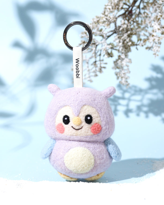 Cute purple owl wool keychain standing on white sand with artistic leaf shadows, designed in Austin as a sustainable slow-fashion accessory