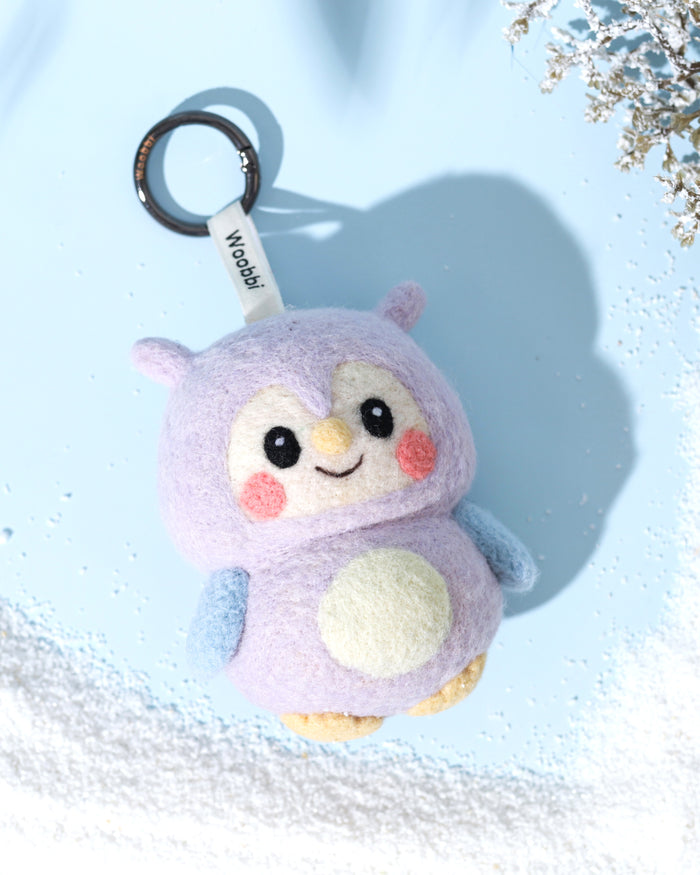 Front view of handcrafted needle-felted wool owl bag charm in purple and blue, featuring a cute smiling face and white branded Woobbi tag, styled on a winter blue background.